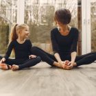 Anonymous mother in sportswear sitting near smiling little blond daughter with crossed legs on parquet while looking at each other in daylight